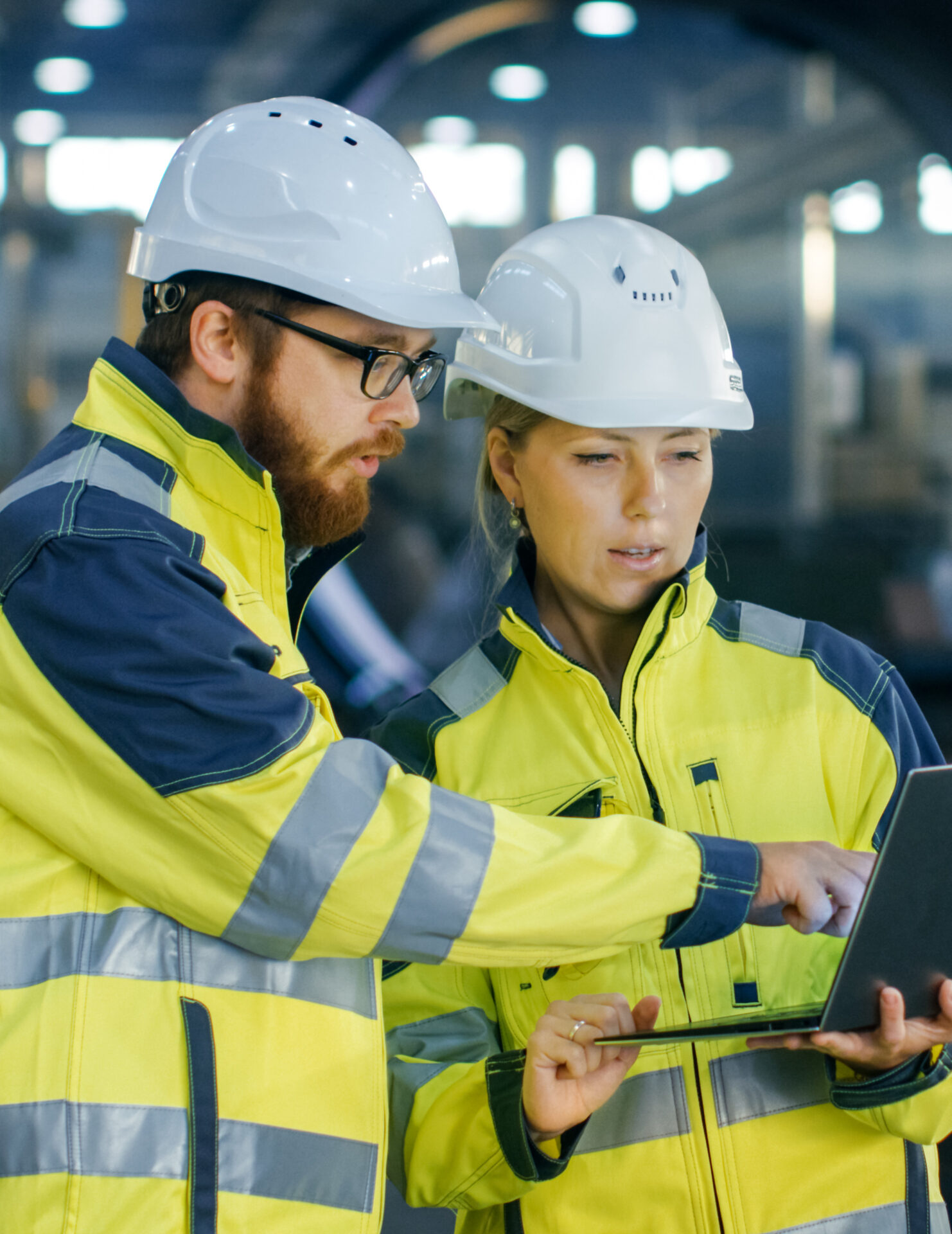 Male and Female Industrial Engineers in Hard Hats Discuss New Project while Using Laptop. They Make Showing Gestures.They Work in a Heavy Industry Manufacturing Factory.